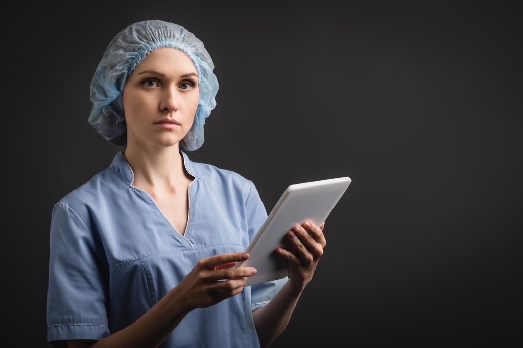 pielęgniarka w czepku medycznym i jednolite trzymanie tabletki cyfrowej odizolowanej na ciemnoszarym - Zdjęcie, obraz