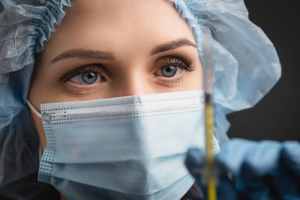 Tıbbi maskeli hemşirenin ön plandaki bulanık aşıyla dolu şırıngayı tutarken koyu gri renkte izole edilmiş. - Fotoğraf, Görsel