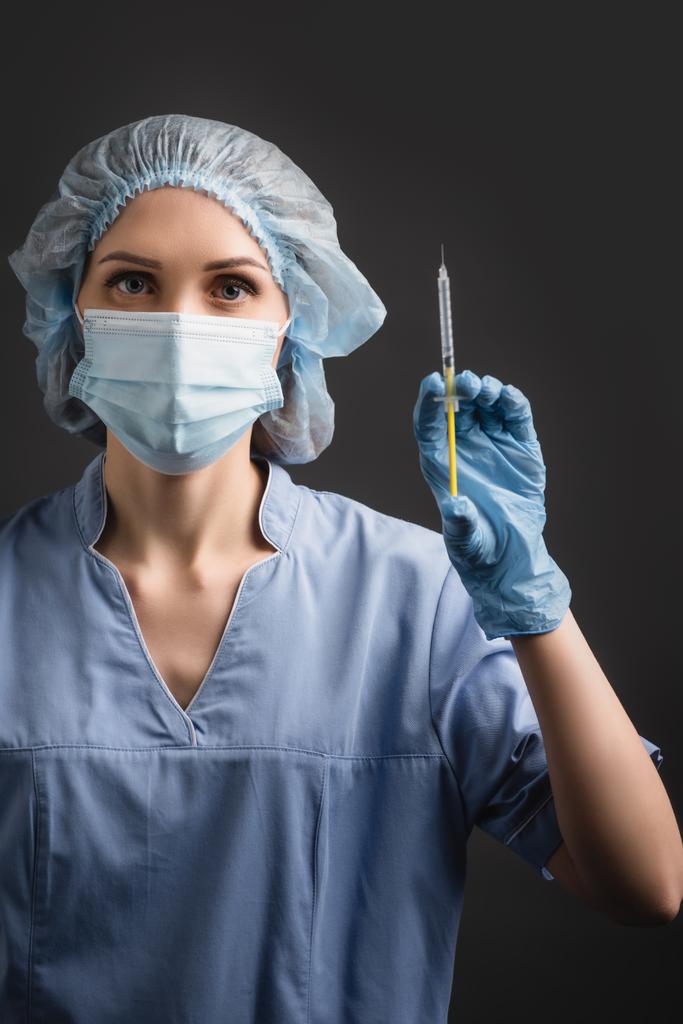 νοσοκόμα με ιατρική μάσκα που κρατά σύριγγα με εμβόλιο απομονωμένο σε σκούρο γκρι - Φωτογραφία, εικόνα