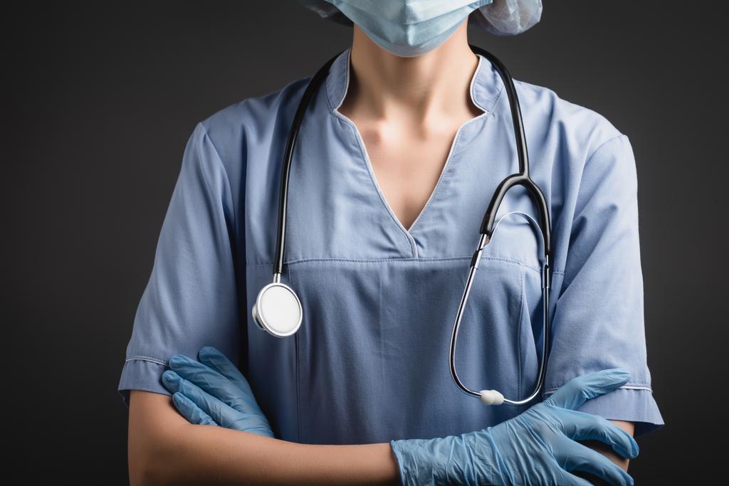 Tıbbi maskeli ve lateks eldivenli hemşirenin kolları çapraz, koyu gri renkte duruyor.  - Fotoğraf, Görsel