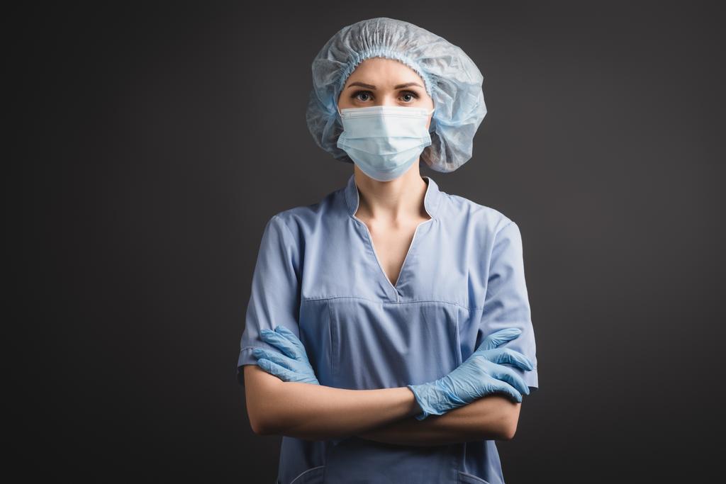 verpleegster in latex handschoenen, medische pet en masker staan met gekruiste armen geïsoleerd op donkergrijs  - Foto, afbeelding