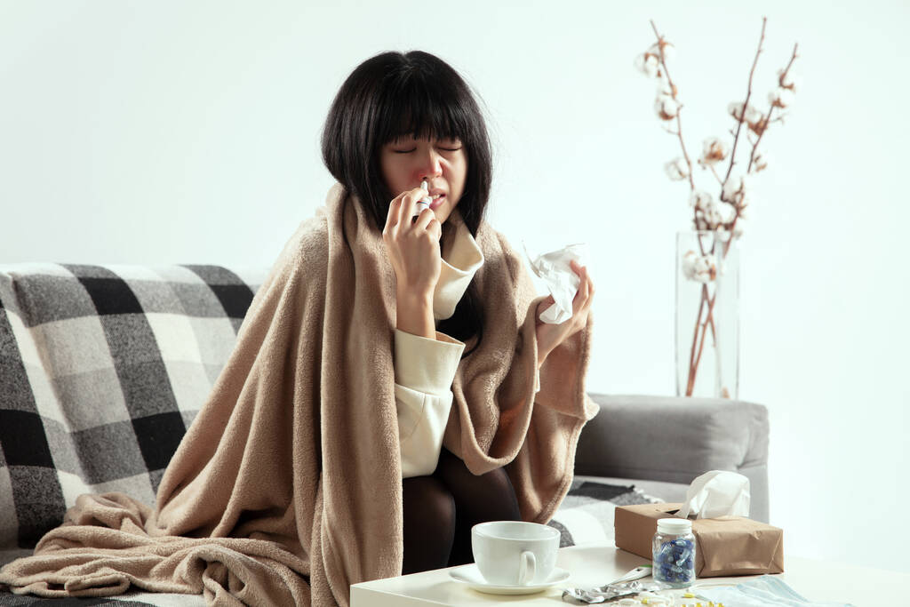 Mujer envuelta en una tela a cuadros se ve enferma, enferma, estornudando y tosiendo sentada en casa dentro - Foto, imagen