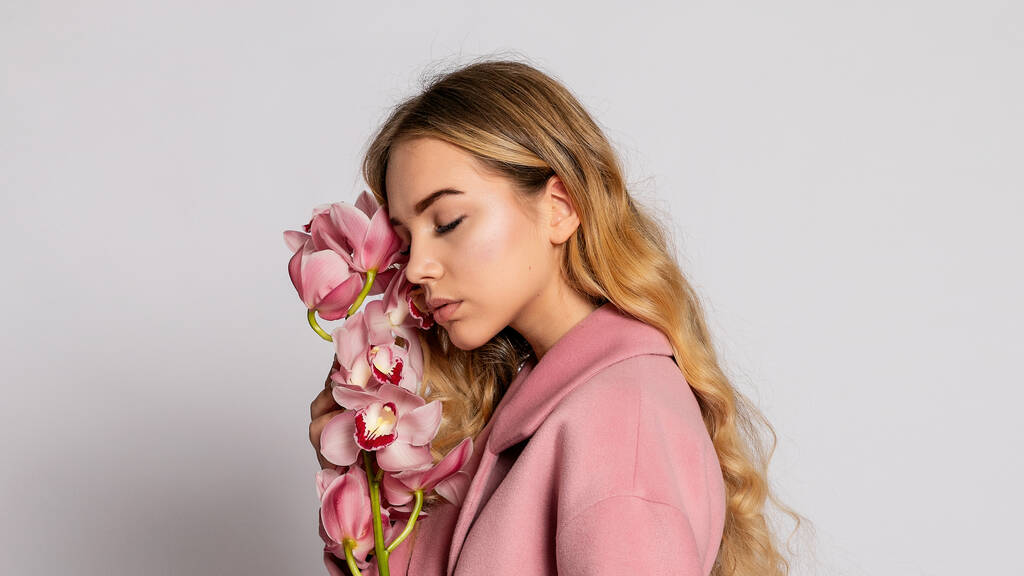 Csábító szőke nő rózsaszín kabátban, szürke háttérrel pózol a stúdióban. elegáns modell pasztell alkalmi tavaszi ruhát. Gyönyörű lány egészséges bőrrel és orchideával. 16: 9 panoráma formátum. - Fotó, kép