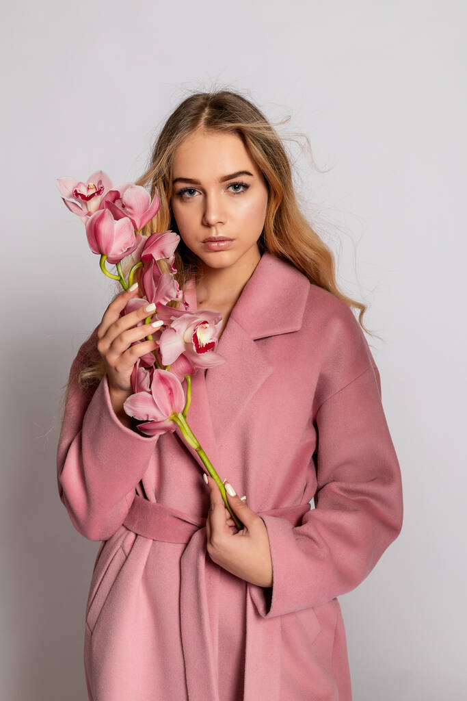 Mujer rubia seductora en chaqueta rosa posando en estudio sobre fondo gris. Retrato de moda de modelo elegante en atuendo de primavera casual pastel. Hermosa chica con la piel sana y orquídea rama - Foto, imagen
