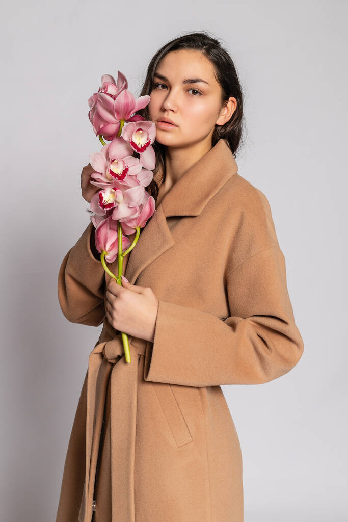 Csábító szőke nő barna kabátban, szürke háttérrel pózol a stúdióban. Divat portré elegáns modell pasztell alkalmi tavaszi ruhát. Gyönyörű lány egészséges bőr és ág orchidea - Fotó, kép