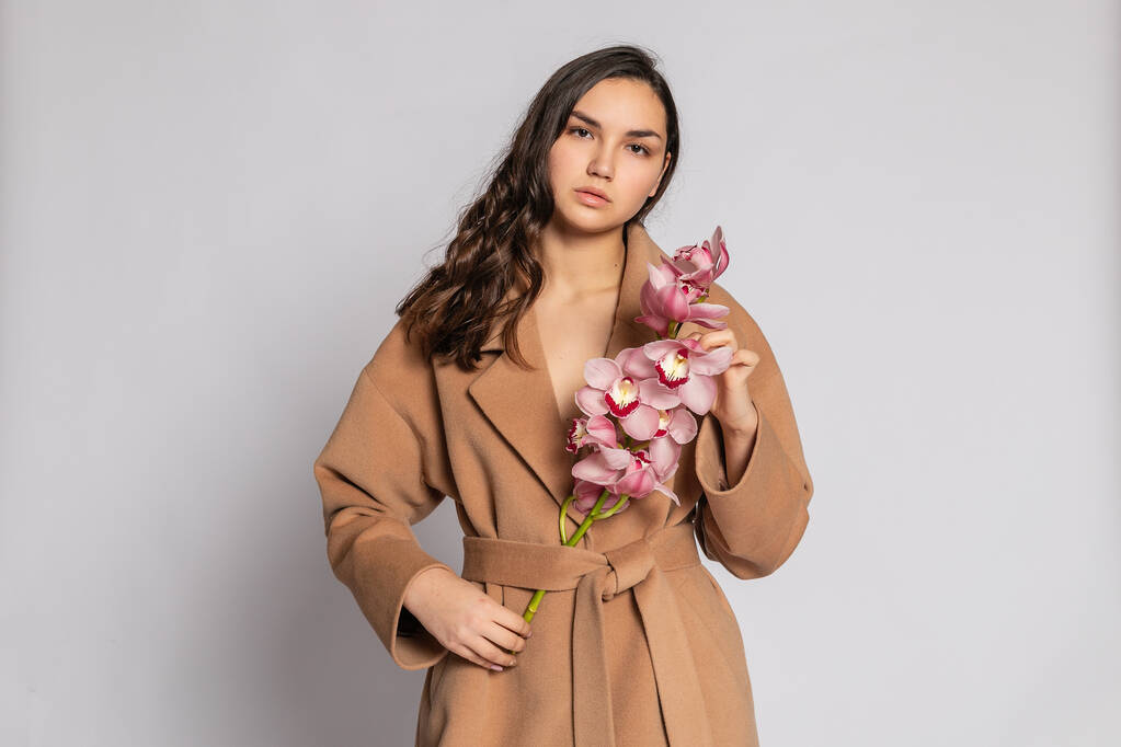 Seductive blonde woman in brown jacket posing in studio on grey background.elegant model in pastel casual spring outfit. Beautiful girl with healthy skin and branch orchid. 16:9 panoramic format. - Photo, Image