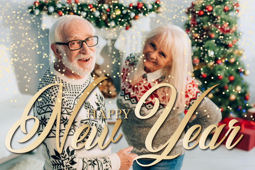 веселый пожилой мужчина и женщина, глядя в камеру рядом с счастливым новогодним письмом и украшения на размытом фоне  - Фото, изображение
