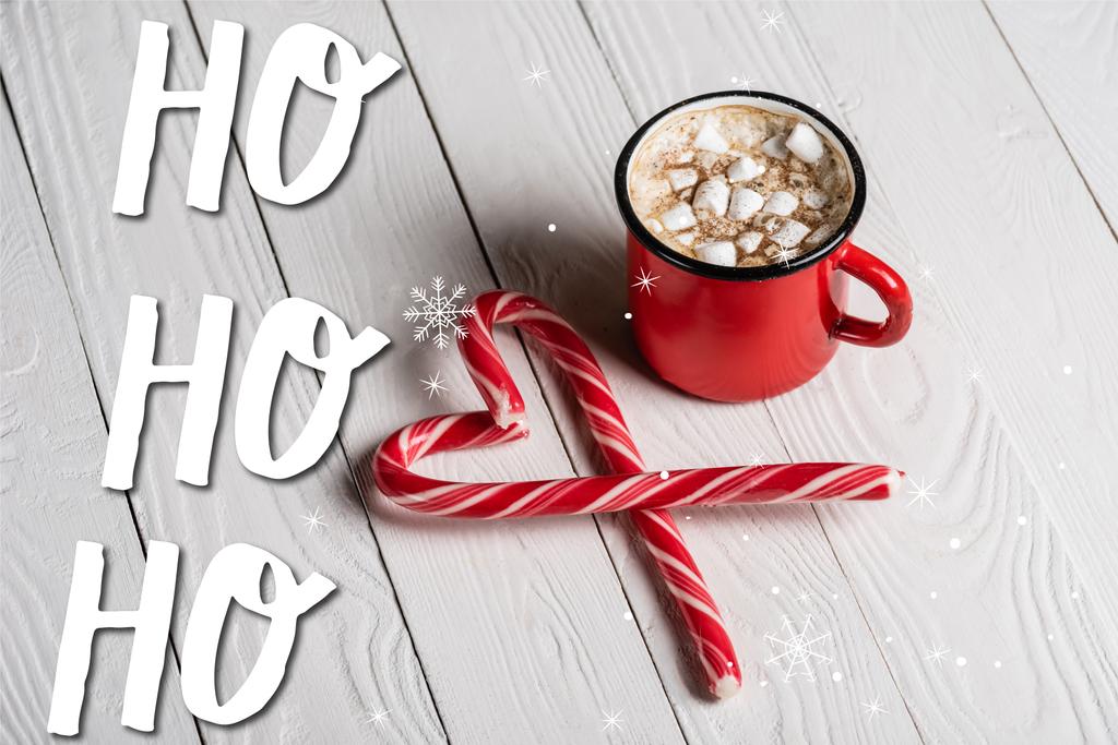Red cup of cocoa near christmas candy canes near  ho ho ho lettering on wooden background - Photo, Image