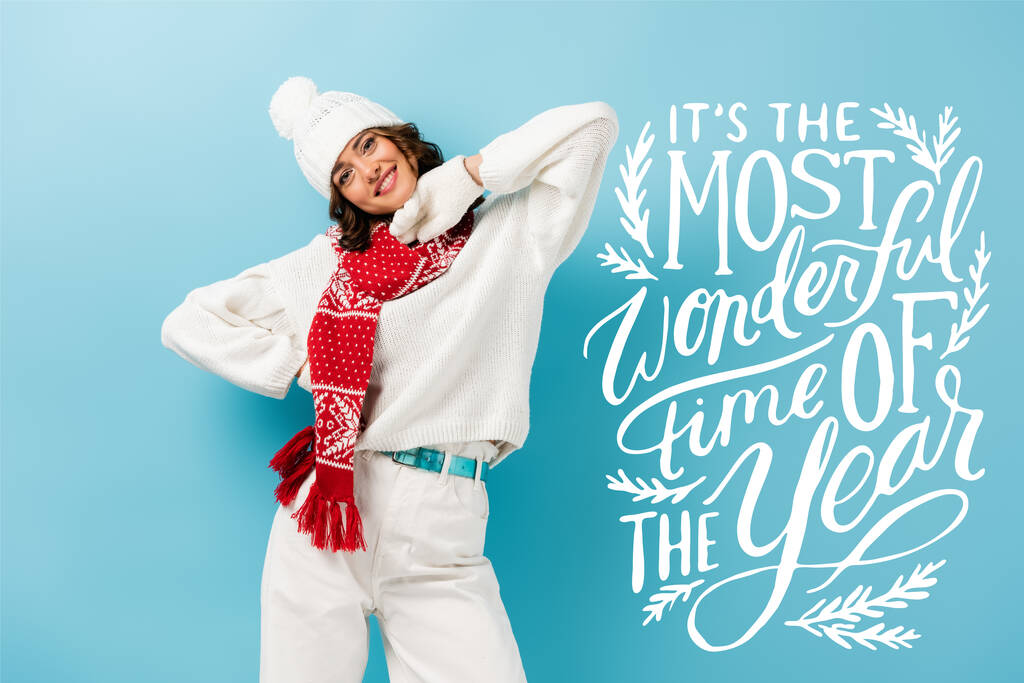 ευχαριστημένη νεαρή γυναίκα σε λευκό χειμωνιάτικο ντύσιμο, ζεστό κασκόλ, γάντια και καπέλο στέκεται κοντά του την πιο υπέροχη εποχή του έτους γράμματα στο μπλε  - Φωτογραφία, εικόνα