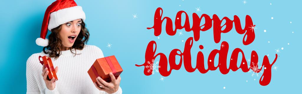 young shocked woman looking at gift box near happy holidays lettering on blue, banner - Photo, Image