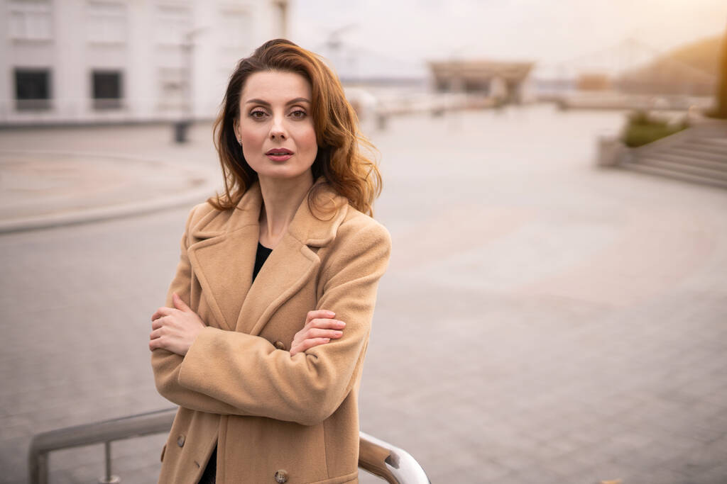 Affascinante giovane donna in un cappotto beige autunno in piedi appoggiato con le braccia incrociate con sfondo urbano della città. Foto tonica - Foto, immagini