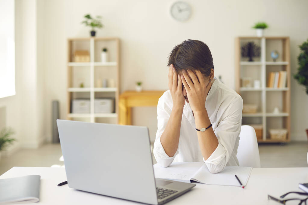 Přepracovaný ženský manažer zavírá tvář unavenýma rukama, sedící před laptopem ve světlé kanceláři. - Fotografie, Obrázek