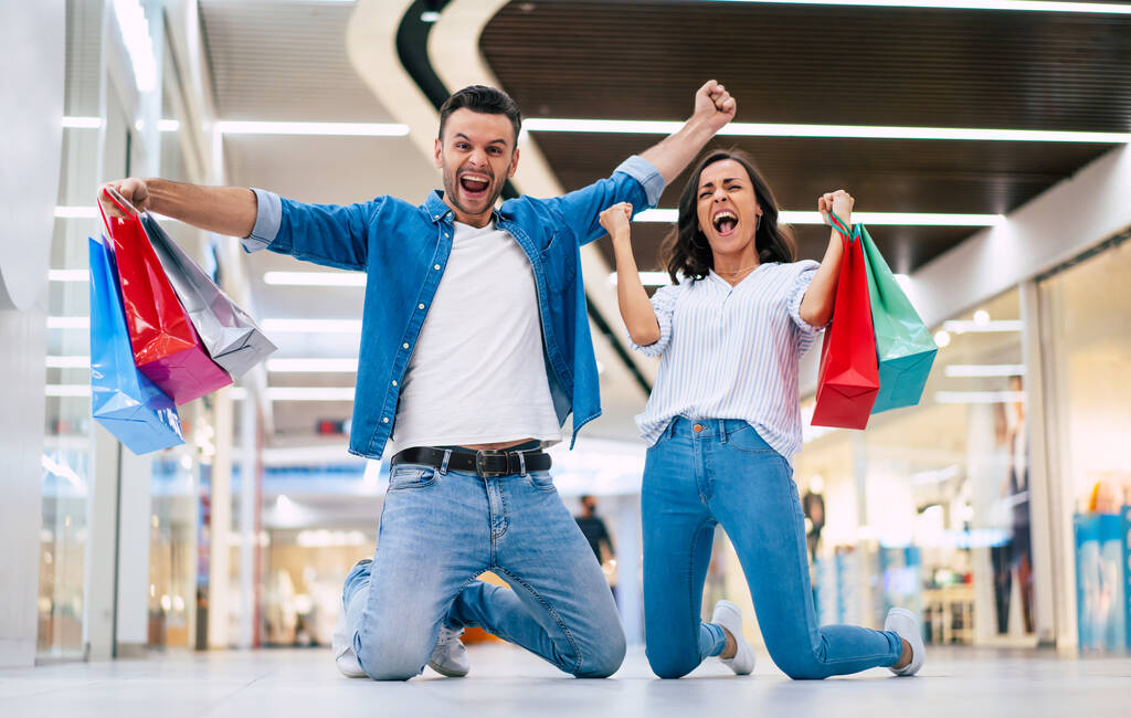  Ενθουσιασμένο ζευγάρι που κερδίζει στην αγάπη ή την οικογένεια με χάρτινες σακούλες στα χέρια ενώ φωνάζουν κατά τη διάρκεια των αγορών στο εμπορικό κέντρο - Φωτογραφία, εικόνα