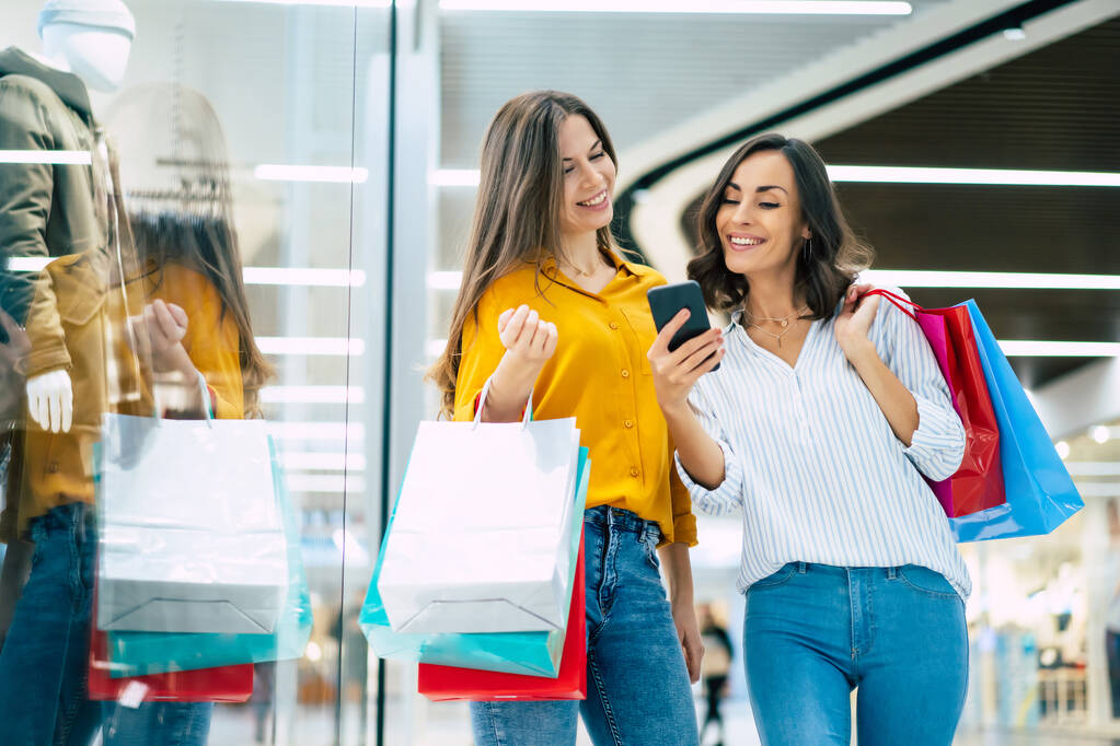Belles amies heureuses et excitées avec des sacs en papier et un téléphone intelligent marchent autour du centre commercial - Photo, image