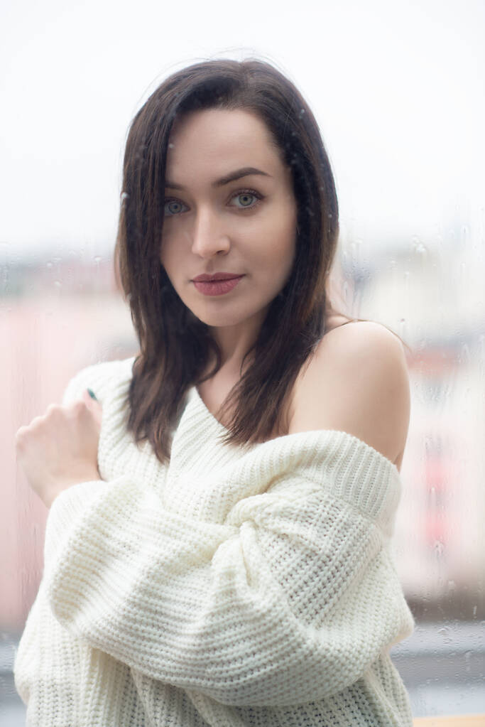 Junge schöne Frau mit langen dunklen Haaren in weißem Pullover, draußen hinter der Fensterscheibe, auf die Regentropfen fallen - Foto, Bild