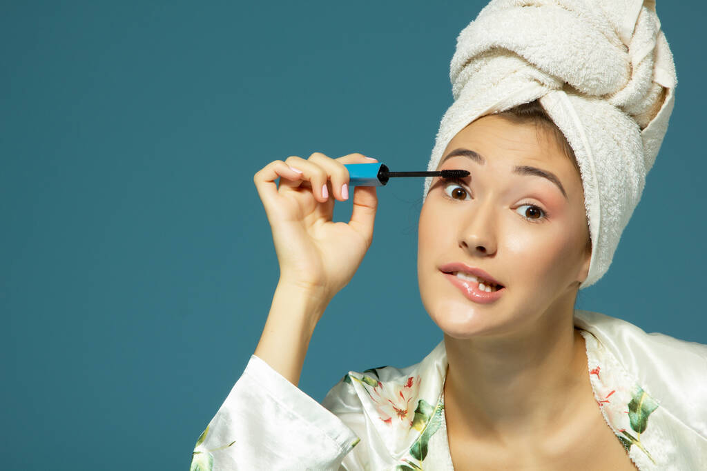 νέα ελκυστική γυναίκα βάζοντας μάσκαρα ματιών πάνω από το μπλε φόντο, θεραπεία ομορφιάς πρωί  - Φωτογραφία, εικόνα