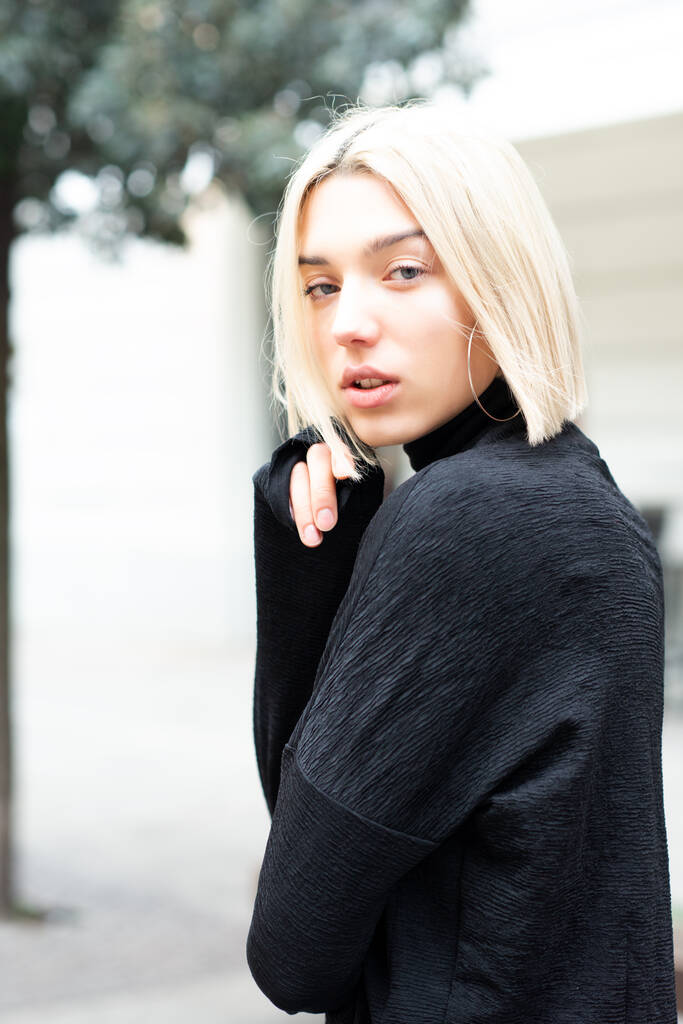Outdoor urban female portrait, soft focus, high key. Fashion model. Young woman posing in Milan streets. Beautiful caucasian girl with blond hair wearing black sweater walking in city. - Photo, Image