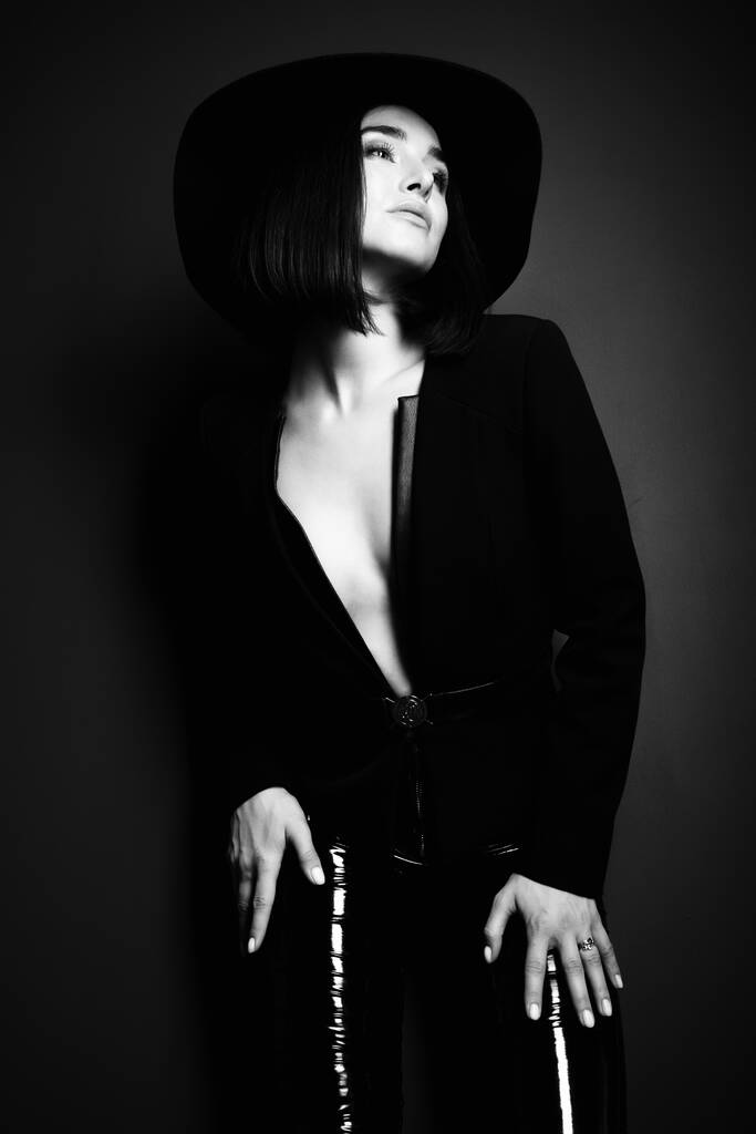 Красивая женщина моды с идеальным телом. Портрет красивой девушки в черном блейзере, латексных узких штанах и шляпе, позирующей в студии на глубоком сером фоне, черно-белом - Фото, изображение