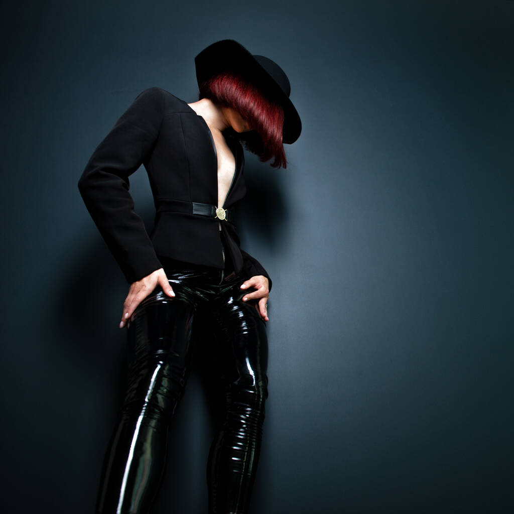 Schöne Modefrau mit perfektem Körper. Porträt eines schönen Mädchens mit schwarzem Blazer, enger Latexhose und Hut, das im Studio vor tiefgrauem Hintergrund posiert - Foto, Bild