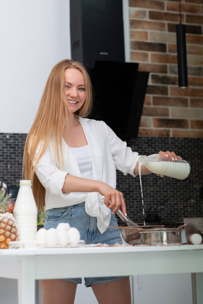 Fiatal nő áll a konyhában otthon, és főzés élvezet pékáru liszt, tej, kakaó, cukor és tojás. Vicces portré boldog lány séf  - Fotó, kép