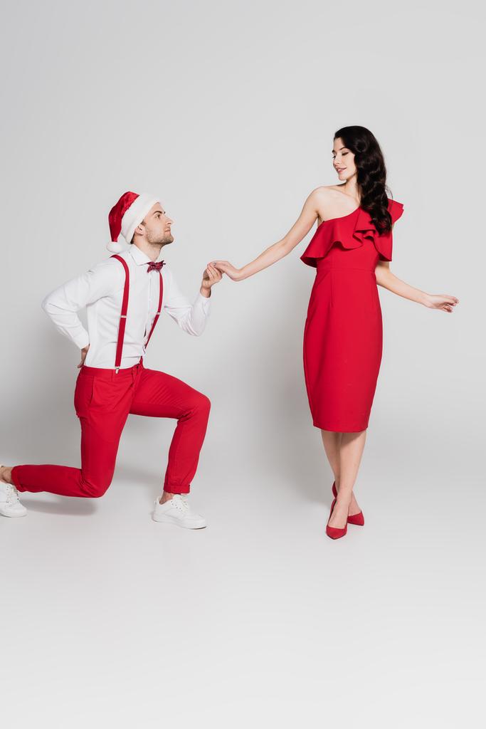 Mann mit Weihnachtsmannmütze steht auf einem Knie und hält Hand einer lächelnden Frau in rotem Kleid auf grauem Hintergrund  - Foto, Bild