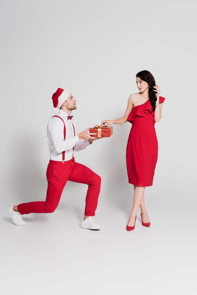 Férfi télapó kalap gazdaság ajándék, miközben áll az egyik térde közelében barátnője piros ruhában szürke háttér  - Fotó, kép