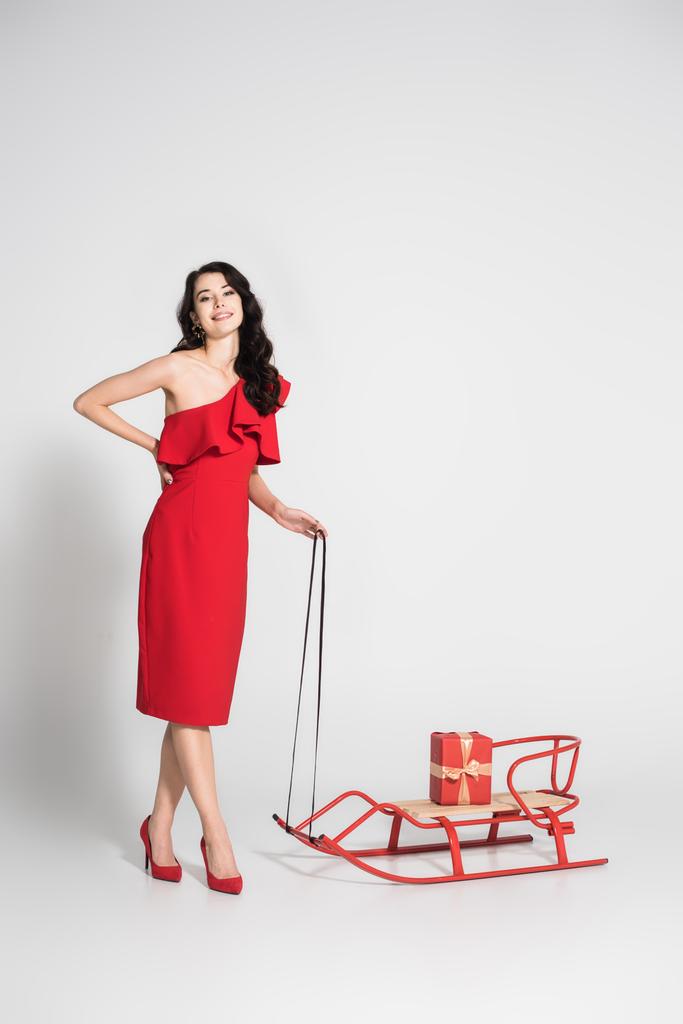 Улыбающаяся брюнетка в красном платье, стоящая возле саней с подарком на сером фоне - Фото, изображение