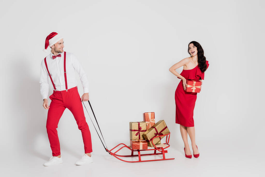 Lächelnder Mann mit Weihnachtsmütze hält Schlitten mit Geschenken in der Nähe einer Frau in rotem Kleid auf grauem Hintergrund  - Foto, Bild