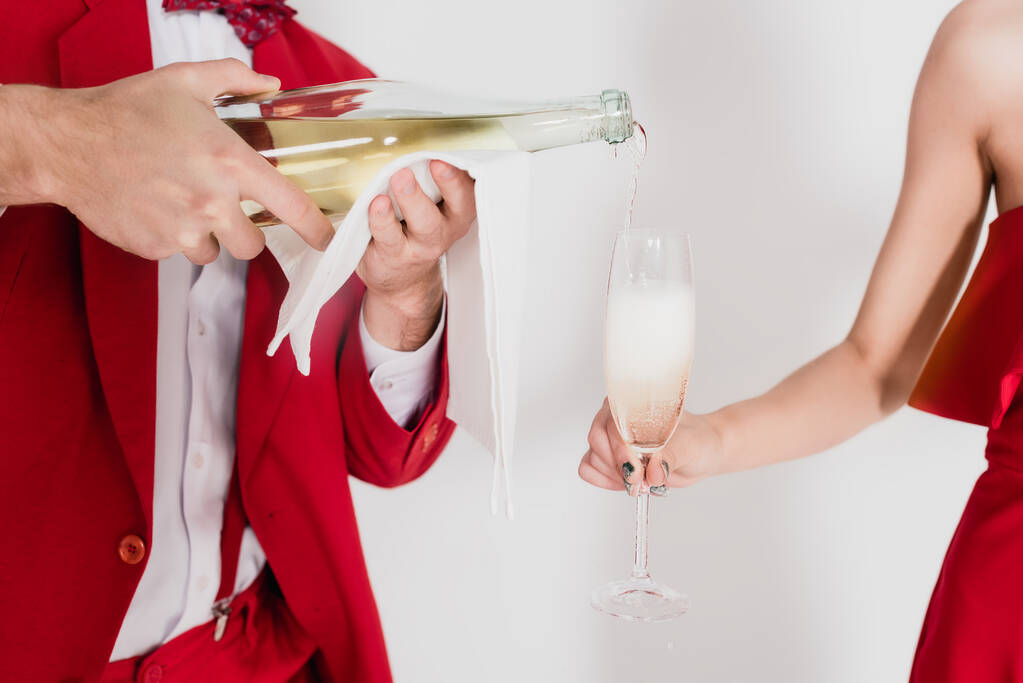 Ausgeschnittene Ansicht eines Mannes im roten Anzug, der Champagner im Glas neben einer Frau auf grauem Hintergrund einschenkt  - Foto, Bild