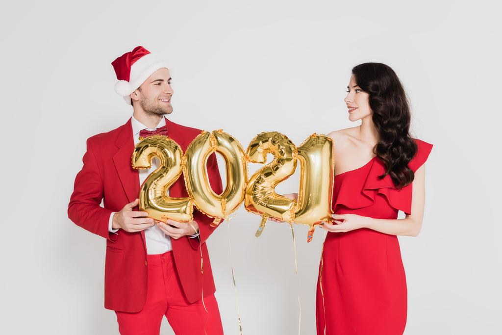 Lächelnder Mann mit Weihnachtsmütze und Frau im roten Kleid mit Luftballons in Form von Ziffern auf grau  - Foto, Bild