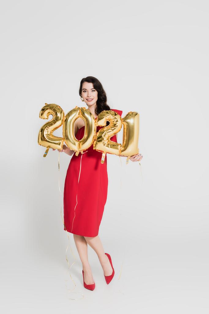 Kırmızı elbiseli neşeli kadın gri arka planda 2021 numaralı balonları tutuyor. - Fotoğraf, Görsel