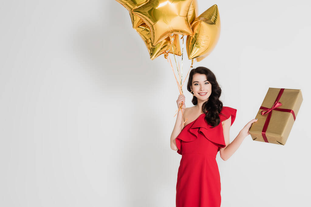 Smiling woman holding balloons and gift box on grey background - Photo, Image