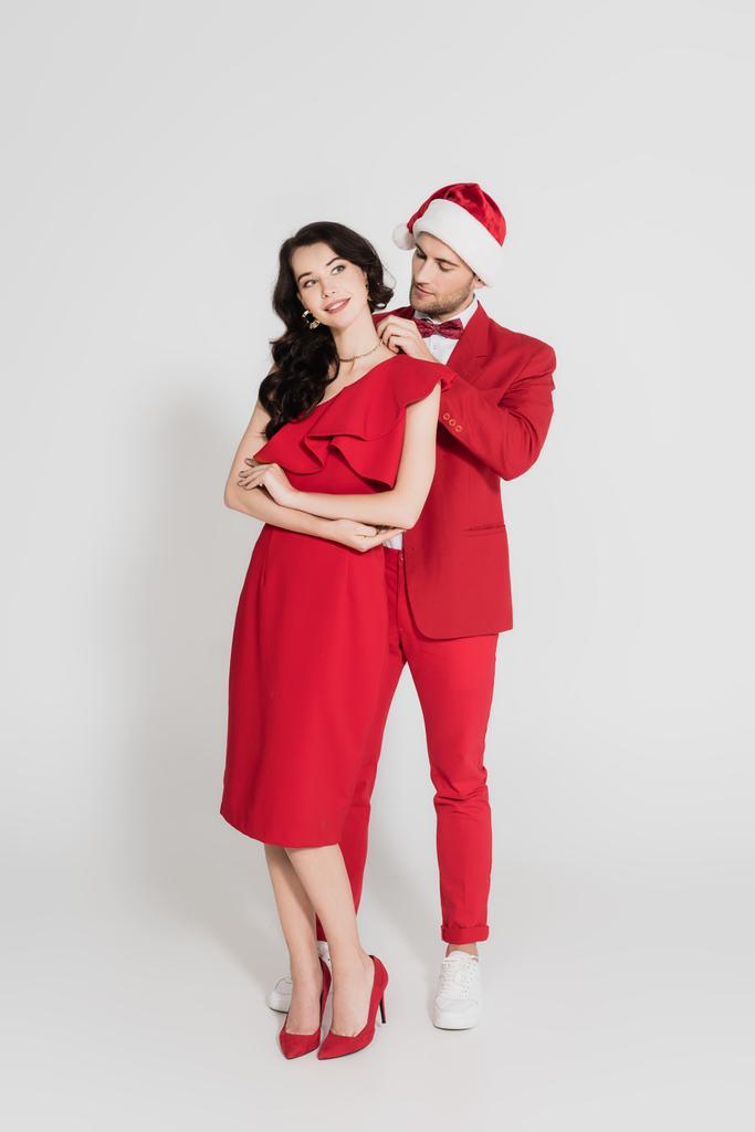 Takım elbiseli ve Noel Baba şapkalı bir adam gri arka planda gülümseyen esmer kadına kolye takıyor.  - Fotoğraf, Görsel