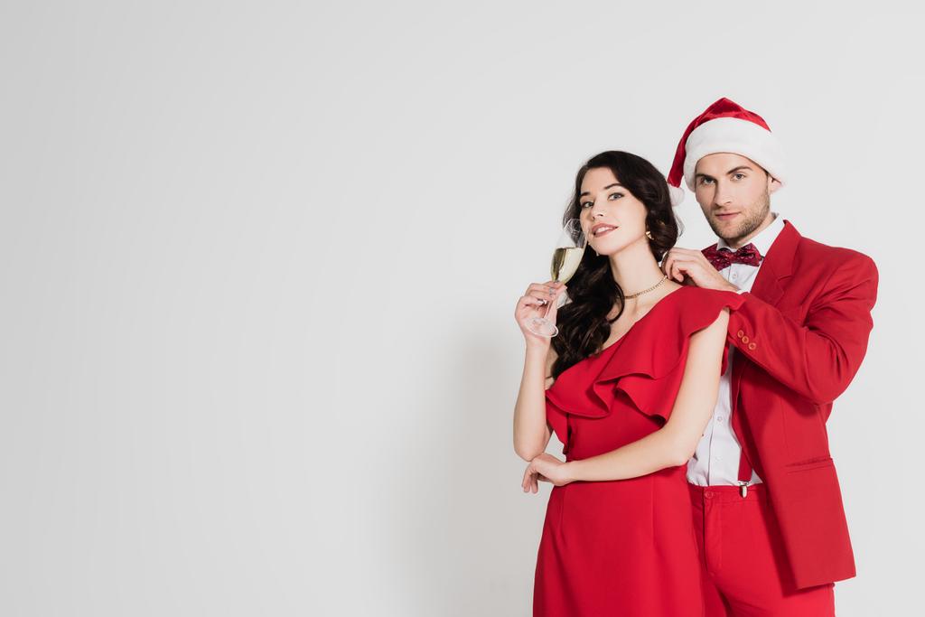 Noel Baba şapkalı ve takım elbiseli adam gri arka planda bir bardak şampanyası olan esmer kadına kolye takıyor.  - Fotoğraf, Görsel