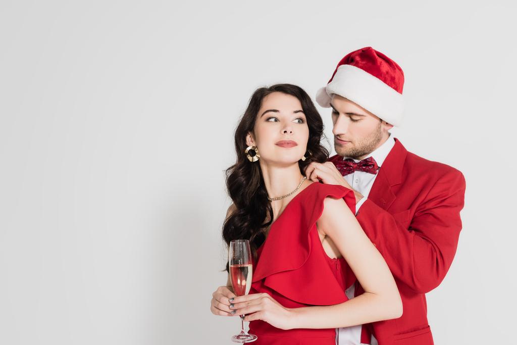 Mann mit Weihnachtsmütze und Fliege trägt Halskette auf Frau im Kleid mit Glas Champagner auf grauem Hintergrund  - Foto, Bild