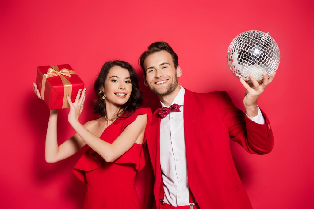 Couple souriant avec cadeau et boule disco regardant la caméra sur fond rouge  - Photo, image