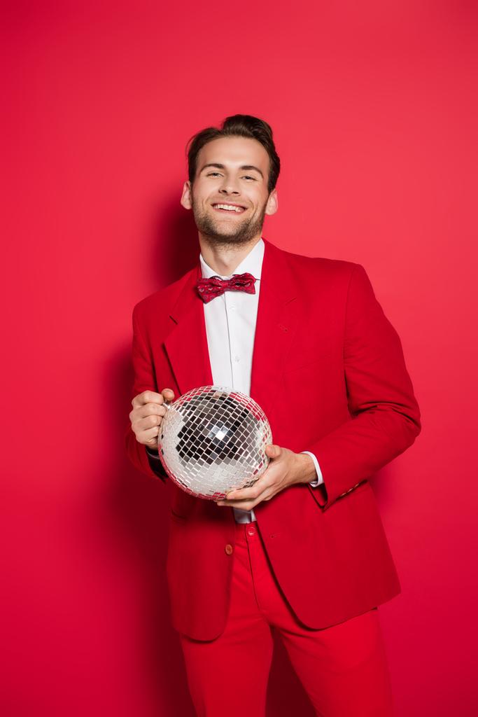 Χαμογελώντας άνθρωπος με κόκκινο κοστούμι κρατώντας ντίσκο μπάλα σε κόκκινο φόντο  - Φωτογραφία, εικόνα