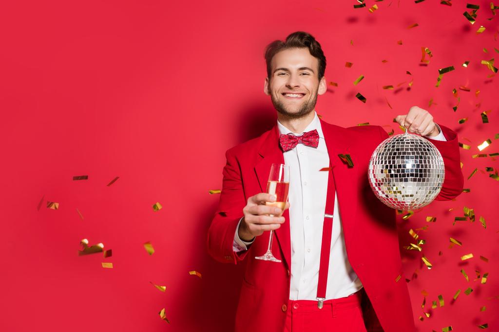 Homme élégant en costume souriant à la caméra tout en tenant boule disco et verre de champagne sur fond rouge  - Photo, image