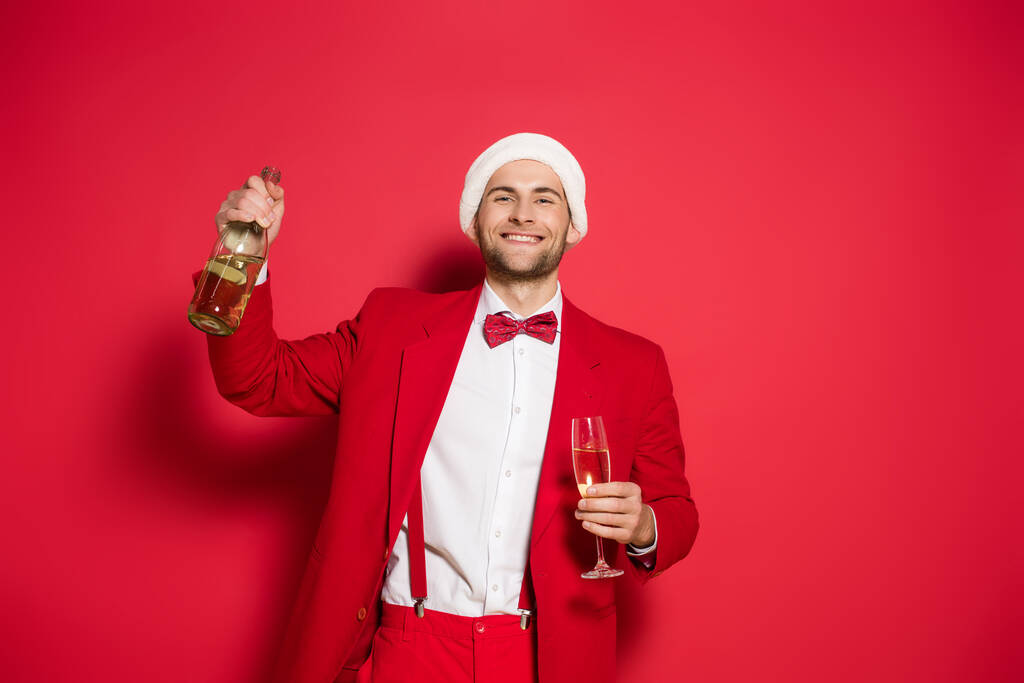 Mladý muž v Santa klobouk a oblek s úsměvem na kameru, zatímco drží láhev a sklenici šampaňského na červeném pozadí  - Fotografie, Obrázek