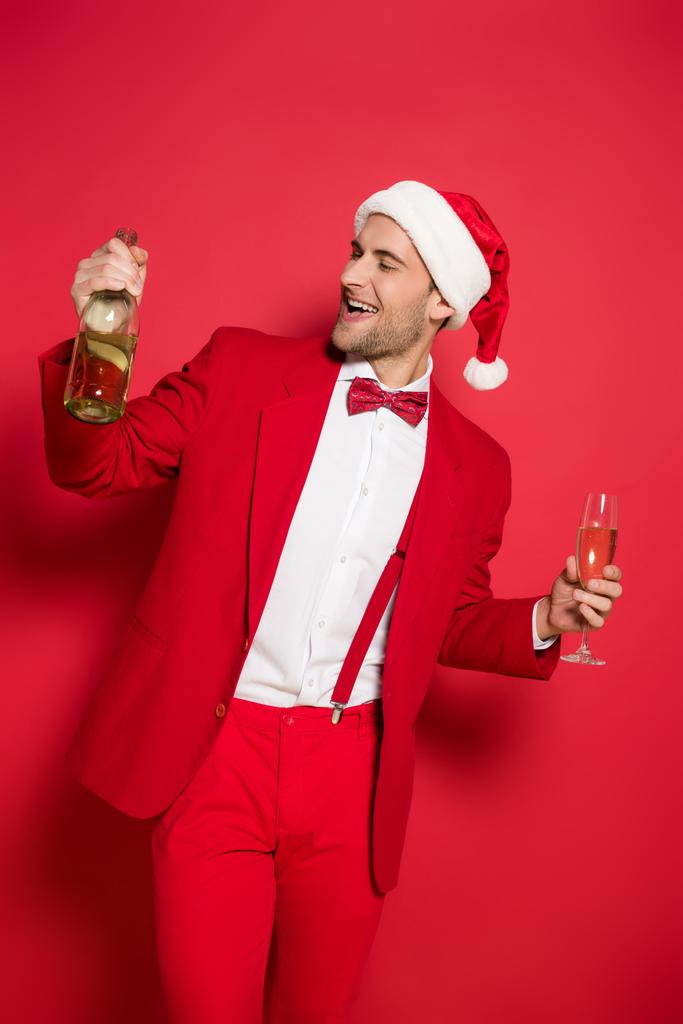 Joven con sombrero de santa y traje rojo sosteniendo botella y copa de champán sobre fondo rojo  - Foto, Imagen