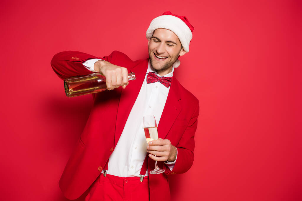 Smiling man in santa hat and suit pouring champagne in glass on red background  - Photo, Image