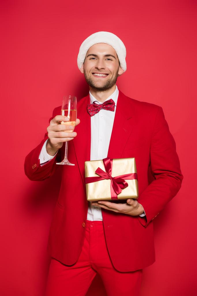 Usmívající se muž v santa klobouk drží dárek a sklenice šampaňského na červeném pozadí  - Fotografie, Obrázek
