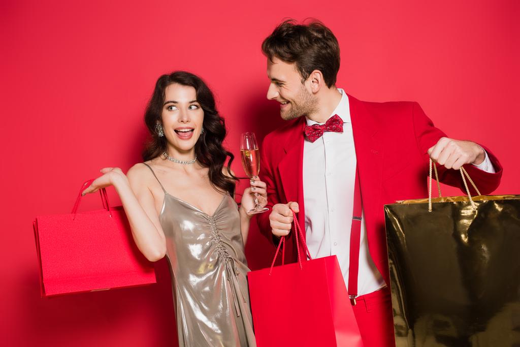 Mujer sonriente sosteniendo copa de champán cerca de novio en traje con bolsas de compras sobre fondo rojo - Foto, imagen