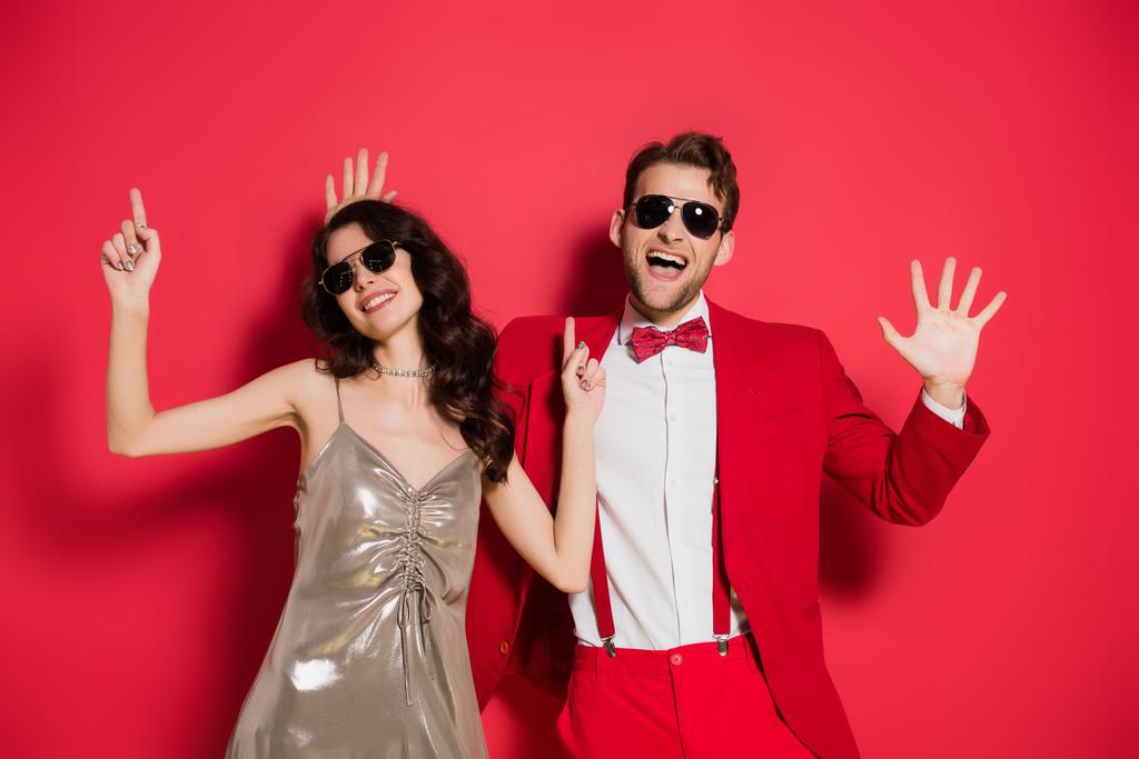 Fröhliches Paar mit Sonnenbrille gestikuliert auf rotem Hintergrund - Foto, Bild