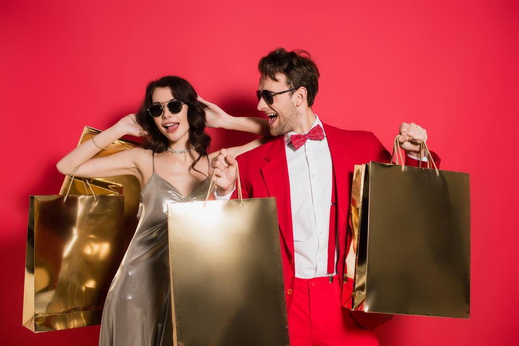 Hombre con estilo en gafas de sol sosteniendo bolsas de compras cerca de novia alegre sobre fondo rojo - Foto, Imagen