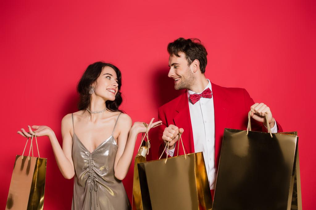 Smiling couple looking at each other while holding shopping bags on red background - Photo, Image