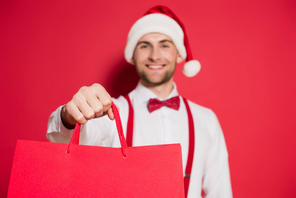 Shopping tassen in de hand van glimlachende man in santa hoed op wazig achtergrond op rode achtergrond - Foto, afbeelding