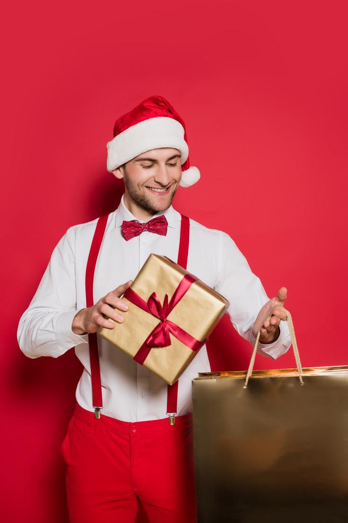 Fröhlicher Mann mit Weihnachtsmütze, Einkaufstasche und Geschenk auf rotem Hintergrund - Foto, Bild