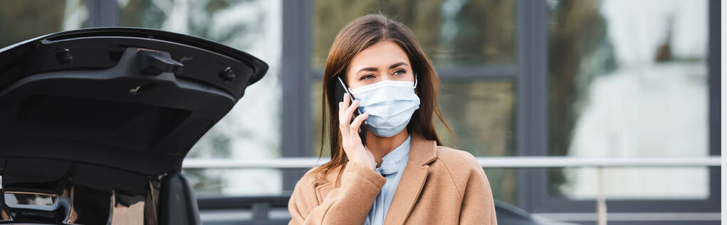mujer joven con máscara médica y traje de otoño hablando en el teléfono inteligente cerca del maletero del coche abierto, pancarta - Foto, Imagen