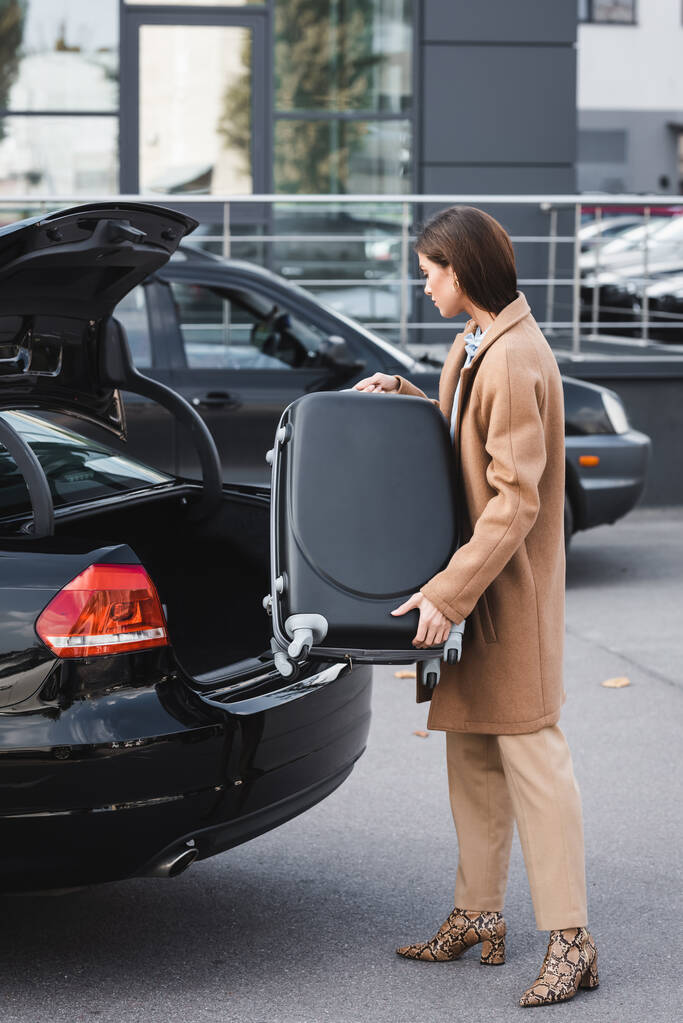 νεαρή γυναίκα με κομψά φθινοπωρινά ρούχα φορτώνει βαλίτσα στο πορτ-μπαγκάζ του αυτοκινήτου - Φωτογραφία, εικόνα